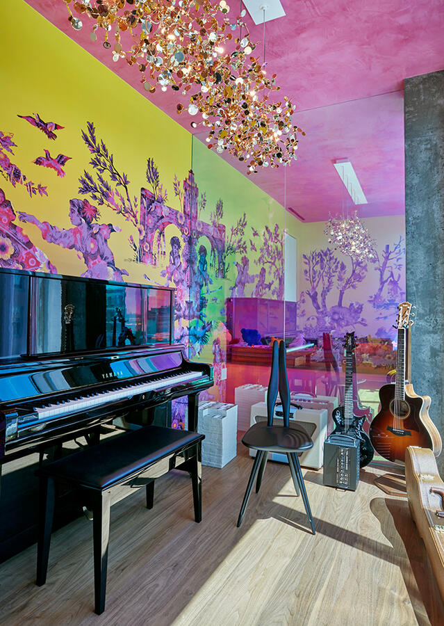A brightly lit room with a colorful wallpaper reflecting off of a mirrored wall. A black piano is flushed against the wallpapered wall, with a bench, stool, and guitars situated around the instrument