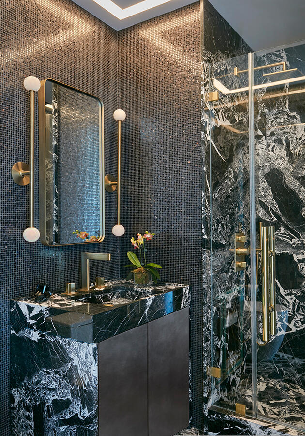 A dark tiled room with dark marble sink and walk-in standing shower with gold accents.