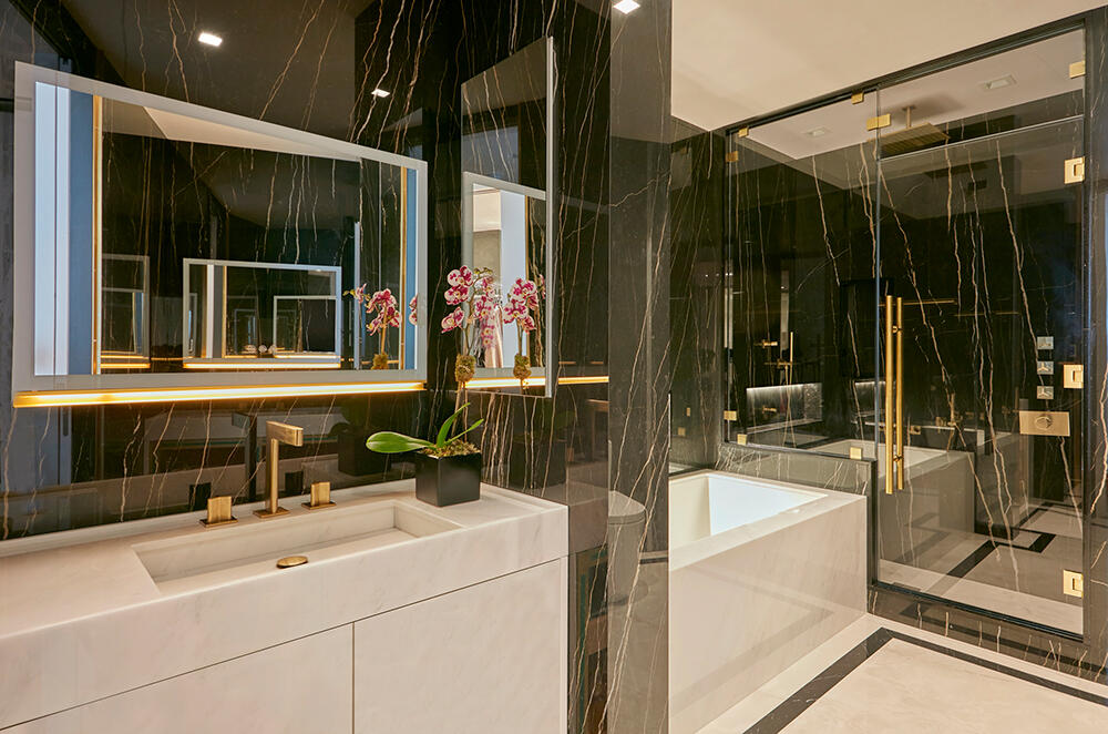 A well lit bathroom with black marble walls. A white quartz sink with gold accented faucet, soaking tub and walk-in shower with a glass separator fills the room.