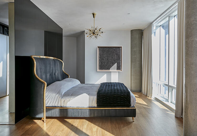 A bedroom with a king sized velvet grey with gold accent bedframe in the middle. A small, decorative chandelier dangles on the wall with sunlight filtering into the room